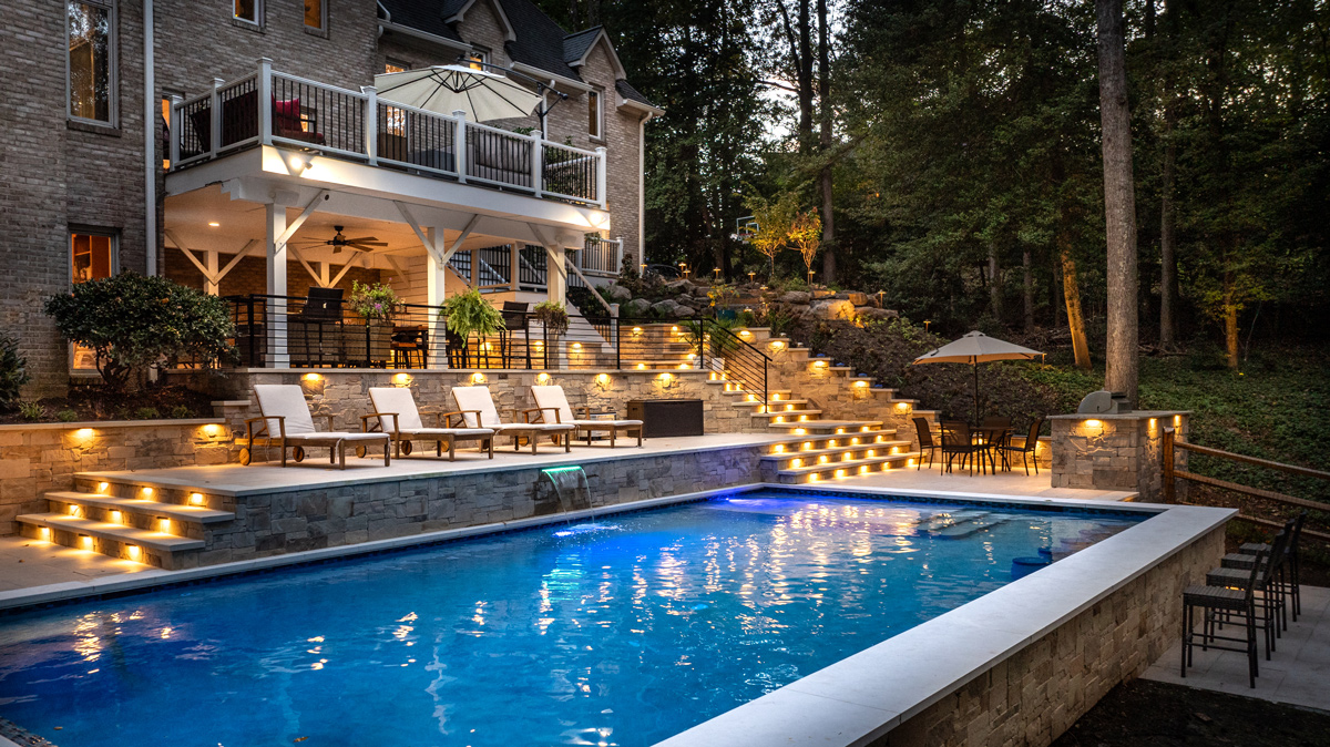 view of the in-ground pool and back patio landscaping lit up at dusk